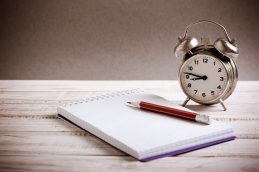Um relógio e um caderno colocados sob uma mesa de madeira, representando a gestão do tempo que precisamos fazer no dia a dia.