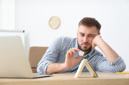 Um homem sentado em uma cadeira, em frente a um notebook e jogando papel em uma agenda, fazendo o que é chamado de procrastinação.