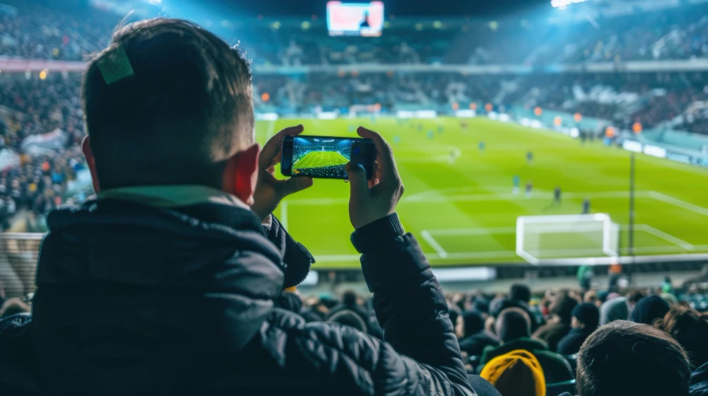 Torcedor assistindo ao jogo de futebol no estádio enquanto grava a partida em telas de smartphone, mostrando a dualidade dos dois cenários.